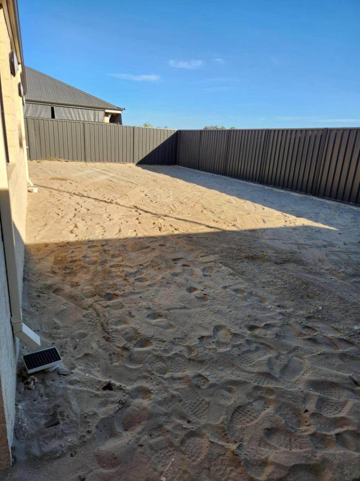 Cleared sandy backyard between a house wall and a steel fence